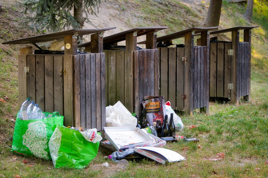 A large collection of overflowing rubbish bins and scattered waste on the pavement in front of a commercial building. The waste includes cardboard boxes, plastic bags filled with various debris, paper